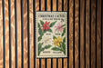 A Christmas cactus postage stamp with the colors red, pink, white, and yellow displayed on it, hanging on a wooden wall.