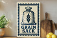 a sign that reads "Grain Sack" with a wheat stalk graphic, placed on a wooden surface next to a wooden cutting board and a potted plant.