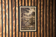 A metal sign with a wood grain background hangs on a wooden wall, featuring an illustration of a gate and trees.