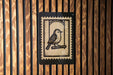 a wooden wall with a black and white postage stamp in the center, depicting a bird perched on a branch.