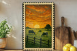 a framed painting depicting a pastoral scene with cows grazing in a grassy field, set against a vibrant sunset sky.