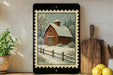 A framed painting of a red barn with a snowy landscape in the background, surrounded by a wooden fence and trees, is displayed on a shelf next to a wooden cutting board and a potted plant.