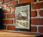 A framed painting of a red barn with a snowy landscape is hanging on a brick wall.