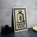 a metal sign with a graphic of a bag of grain and the text "Grain Sack" on it, placed on a countertop next to some black camera lenses.
