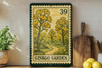 a framed print of a garden scene with a path leading through it, surrounded by yellow flowers and greenery. The print is framed in a green border and has the text "GINKGO GARDEN" printed on it. The print is displayed on a shelf next to a wooden cutting board and a potted plant.