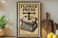 a vintage-style sign for a "flower press" on a wooden shelf, with a potted plant and kitchen utensils visible in the background.