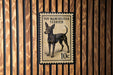 A toy Manchester Terrier dog is depicted on a postage stamp-like sign, mounted on a wooden wall.