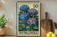 A vintage-style metal stamp depicting a group of blue hydrangea flowers, with the number 50 and the word "HYDRANGEA" printed on it. The stamp is placed on a wooden surface, surrounded by various kitchen utensils and a potted plant.