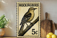 A vintage-style postage stamp featuring a detailed illustration of a bird perched on a branch, with the text "MOCKINGBIRD" and the denomination "5c" printed on it. The stamp is displayed on a shelf next to a wooden cutting board and a potted plant.