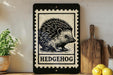 a black and white illustration of a hedgehog on a wooden frame, with the word "HEDGEHOG" written below it. The frame is placed on a wooden surface, with a potted plant to the left and a wooden cutting board to the right.