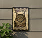 A wooden post with a postage stamp-like design featuring a cat's face, mounted on a wall next to a plant.