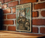 A vintage-style metal stamp depicting a mouse wearing a robe and holding a basket of mice, mounted on a brick wall.