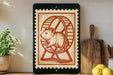 a framed stamp-like artwork depicting a hamster running on a wheel, placed on a shelf next to a wooden cutting board and a potted plant.