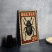 A metal sign featuring a detailed illustration of a beetle, with the word "BEETLE" prominently displayed. The sign is placed on a table, with a stack of black camera lenses and a white plate visible in the background.