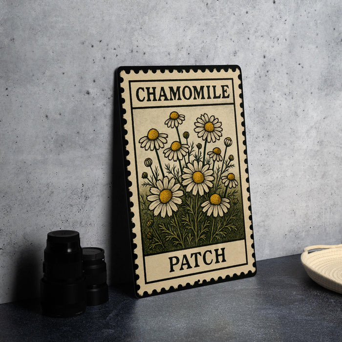 A metal stamp-like object with a floral design and the text "CHAMOMILE PATCH" printed on it, placed on a dark surface next to some black objects.