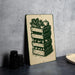 A metal sign with a green and white illustration of a stack of crates containing various vegetables, such as corn, tomatoes, and apples, is displayed on a gray wall.
