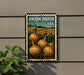 A vintage-style postage stamp featuring an illustration of onions growing in a field, mounted on a wall next to a plant.