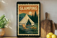 A framed vintage-style poster of a camping scene with a tent, chair, and table, along with a potted plant and wooden cutting board, is displayed on a wall.