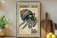 A vintage-style metal sign featuring a hermit crab, with the text "HERMIT CRAB" and "50c" printed on it. The sign is placed on a wooden surface, with a potted plant, a wooden cutting board, and some lemons nearby.
