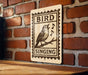A sign with a bird perched on a branch, with the text "BIRD SINGING" below it, mounted on a brick wall.