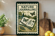 A framed poster with a nature-themed illustration, including a book, butterflies, and plants, is displayed on a shelf next to a wooden cutting board and a potted plant.