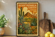 a framed poster of a desert botanical garden with various cacti and succulents, placed on a shelf next to a wooden cutting board and a potted plant.