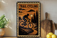 A vintage-style metal sign with a silhouette of a person riding a bicycle, depicting a trail cycling scene, is displayed on a shelf next to a wooden cutting board and some potted plants.