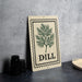 a decorative sign with a tree design and the word "DILL" printed on it, placed on a countertop next to some black containers.
