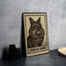 A framed illustration of a lionhead rabbit is displayed on a table, accompanied by a stack of black camera lenses and a white plate.