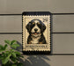 A Bernoodle dog is depicted on a postage stamp-like sign, mounted on a wall next to a plant.