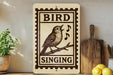 a wooden sign with a bird perched on a branch, singing, and the word "SINGING" written below the bird. The sign is placed on a shelf next to a potted plant and a cutting board.