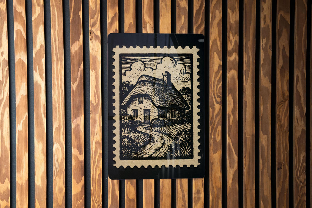 A black and white illustration of a thatched roof cottage is displayed on a wooden wall, with a black border around it.