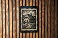 A black and white illustration of a thatched roof cottage is displayed on a wooden wall, with a black border around it.