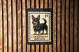 A black and white postage stamp featuring a dog, specifically a Lancashire Heeler, mounted on a wooden wall.