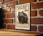 A black bear hamster postage stamp is displayed on a brick wall, with a wooden table below it.