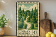A framed illustration of a topiary garden, featuring a winding path surrounded by lush, meticulously trimmed hedges. The garden is depicted in a whimsical, artistic style, with a variety of green trees and bushes. The frame has a vintage or antique appearance, and the text "TOPIARY GARDEN" is visible, indicating the subject of the artwork.