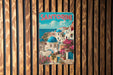 A vintage metal sign with the word "SANTORINI" in large letters, depicting a picturesque coastal town with white buildings, blue domes, and pink flowers.