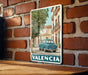 A vintage-style poster of a car parked on a street in a city, with a tree and buildings in the background, mounted on a brick wall.