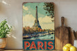A vintage-style metal sign featuring the Eiffel Tower in Paris, with a potted plant to the left and a wooden cutting board to the right.