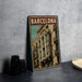 A framed poster of a building in Barcelona, Spain, is displayed on a table next to some black camera lenses.