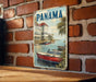 A vintage-style metal sign with the word "PANAMA" prominently displayed, featuring a scenic image of boats on a body of water with a brick wall in the background.