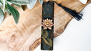 A decorative bookmark featuring a pink lotus flower and green leaves rests on a wooden surface, accompanied by a black tassel.