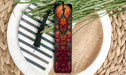 A decorative bookmark with a peacock design is placed on a plate with a striped pattern, surrounded by green foliage.