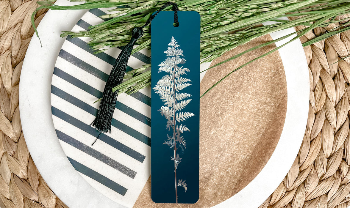 A blue bookmark with a white fern design rests on a white plate with a black and white striped pattern, surrounded by green foliage.