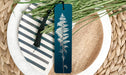 A blue bookmark with a white fern design rests on a white plate with a black and white striped pattern, surrounded by green foliage.