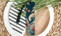 A decorative bookmark with a wave design and a tassel is placed on a plate with a striped pattern and a pile of green grass.