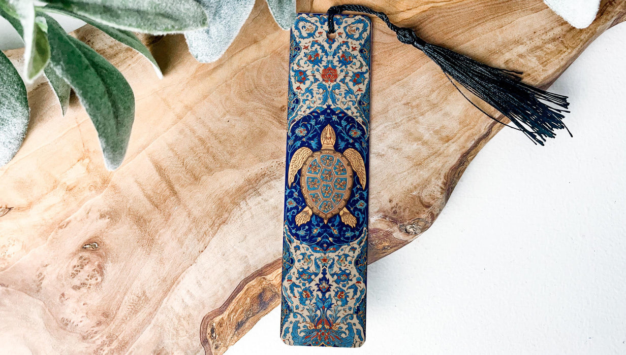 A decorative blue and gold turtle-themed bookmark with a black tassel is placed on a wooden surface, surrounded by green leaves.