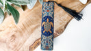A decorative blue and gold turtle-themed bookmark with a black tassel is placed on a wooden surface, surrounded by green leaves.
