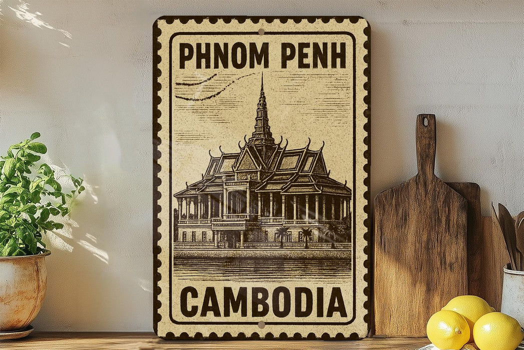 A vintage-style metal sign with the text "PHNOM PENH" and "CAMBODIA" printed on it, along with an illustration of a traditional Thai temple. The sign is displayed on a shelf next to a wooden cutting board and a potted plant.
