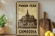 A vintage-style metal sign with the text "PHNOM PENH" and "CAMBODIA" printed on it, along with an illustration of a traditional Thai temple. The sign is displayed on a shelf next to a wooden cutting board and a potted plant.